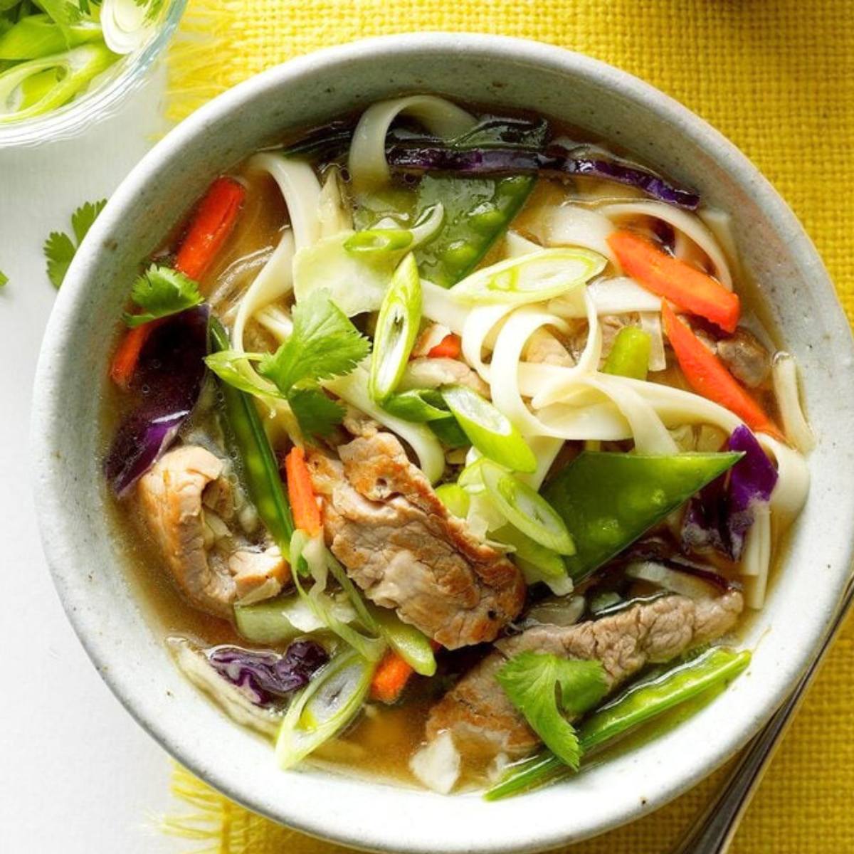 A bowl of noodle soup filled with colorful vegetables like carrots, cabbage, and snow peas. The soup also contains slices of meat and is garnished with fresh herbs.