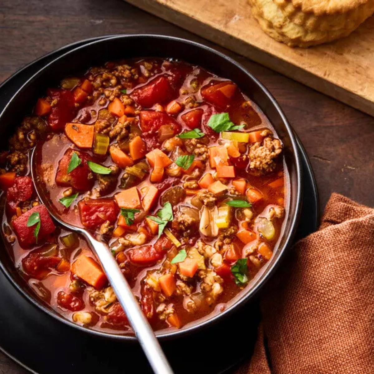 A black bowl filled with a steaming bowl of tomato soup. The soup is made with ground beef, barley, carrots, and celery, and is garnished with fresh parsley. A spoon is resting in the bowl, and a plate of biscuits is visible in the background.