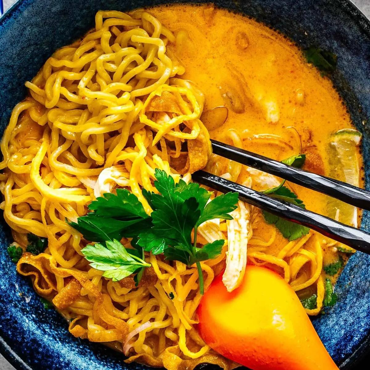 A blue bowl filled with a golden curry soup. Egg noodles are topped with a creamy, coconut-based curry sauce, garnished with fresh herbs and a red chili pepper. Chopsticks are used to lift a strand of noodle.