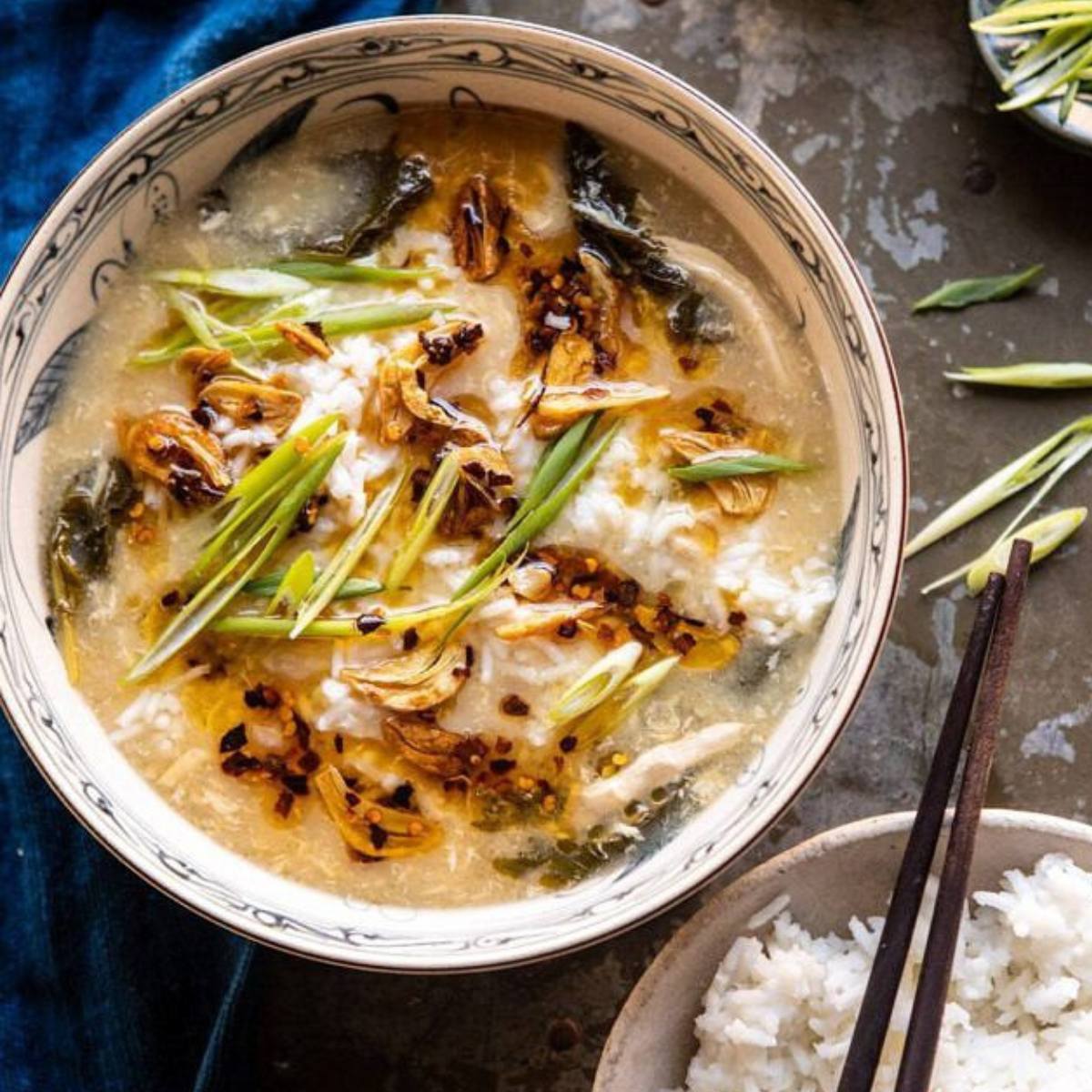 A bowl of creamy soup topped with crispy garlic, green onions, and chili oil. A side of rice and chopsticks are also visible.