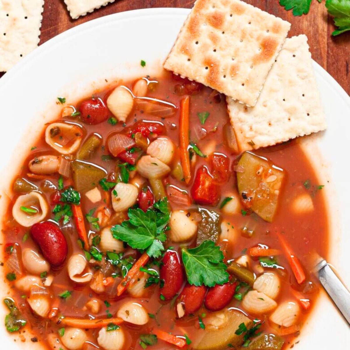 A close-up shot of a white bowl filled with a colorful and flavorful minestrone soup, served with crackers.