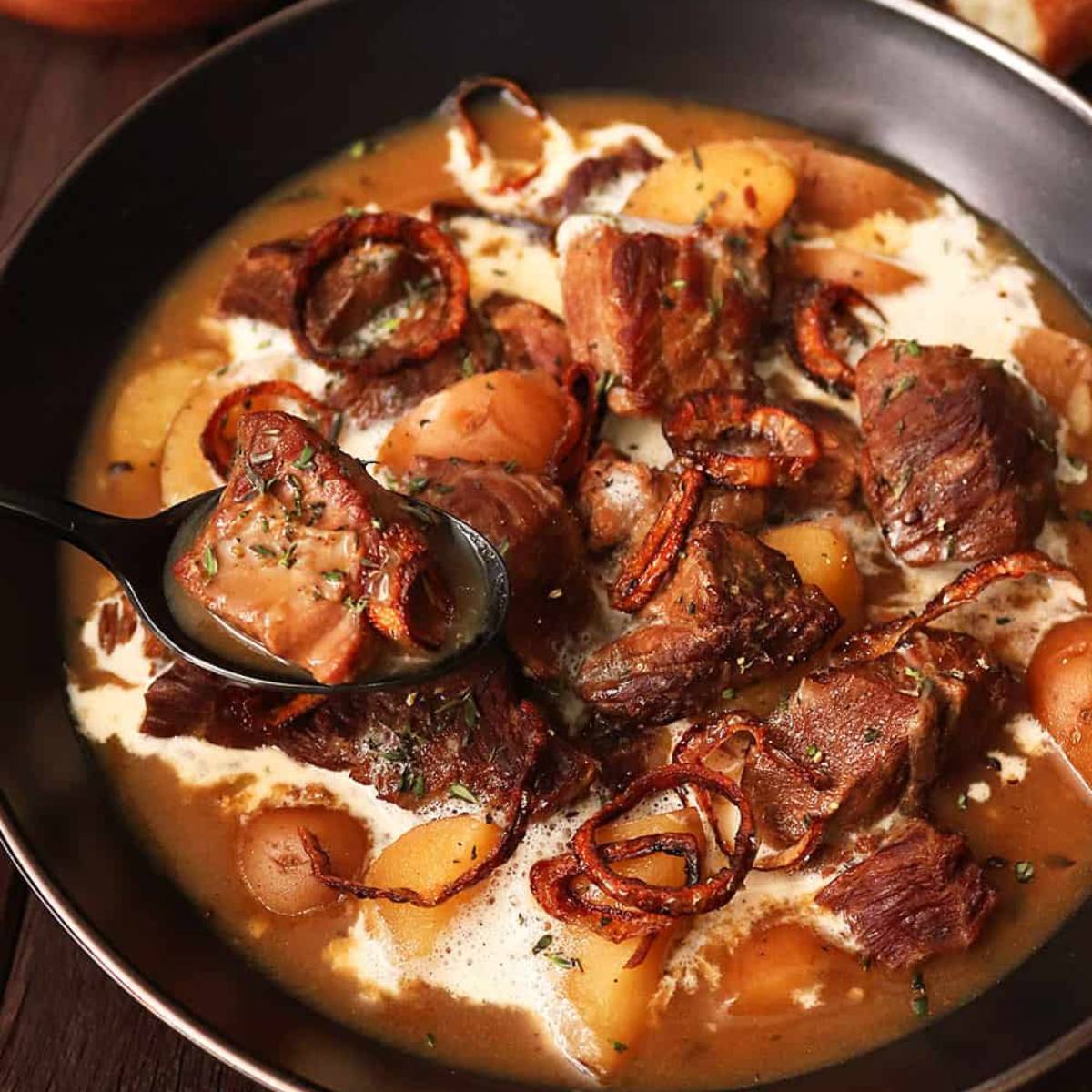 A black bowl filled with a rich, creamy beef stew. Tender chunks of beef, potatoes, and onions are simmered in a flavorful sauce. A spoon is scooping up a bite of the stew.