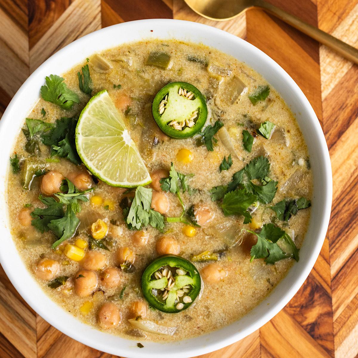 A white bowl filled with a creamy, golden soup. The soup is topped with chickpeas, corn, lime wedges, jalapeno slices, and fresh cilantro.