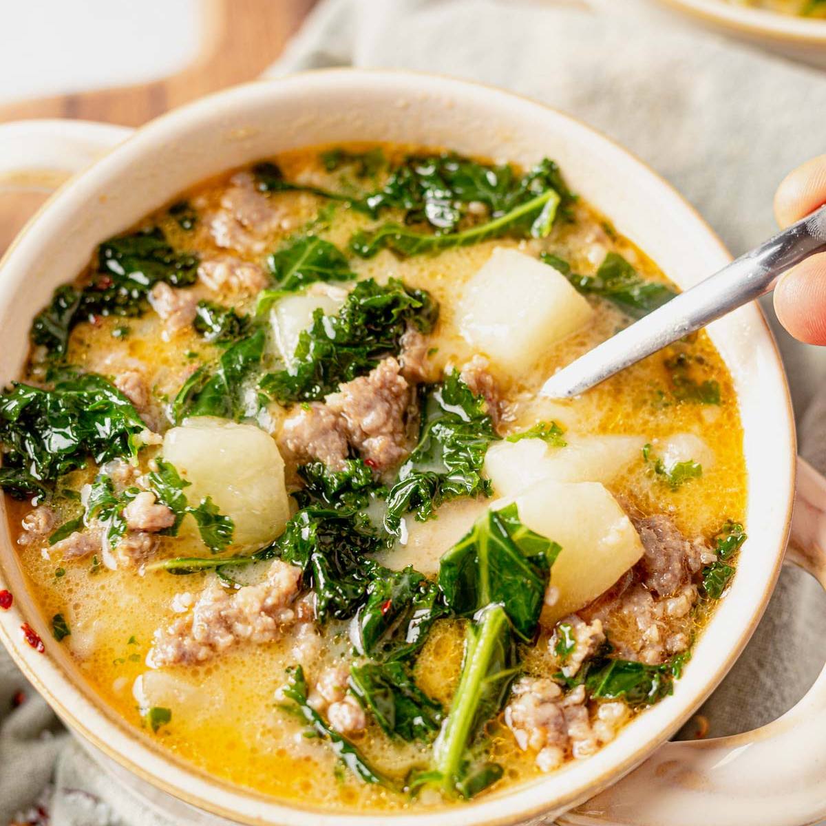 A white bowl filled with a hearty sausage and kale soup. The soup is made with sausage, potatoes, kale, and a creamy broth. A spoon is scooping up a bite of the soup.