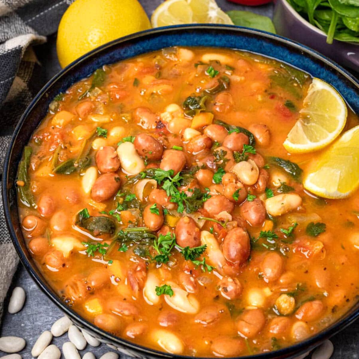 A blue bowl filled with a vibrant bean soup. The soup is made with a variety of beans, vegetables, and herbs, and is garnished with lemon slices and fresh parsley.