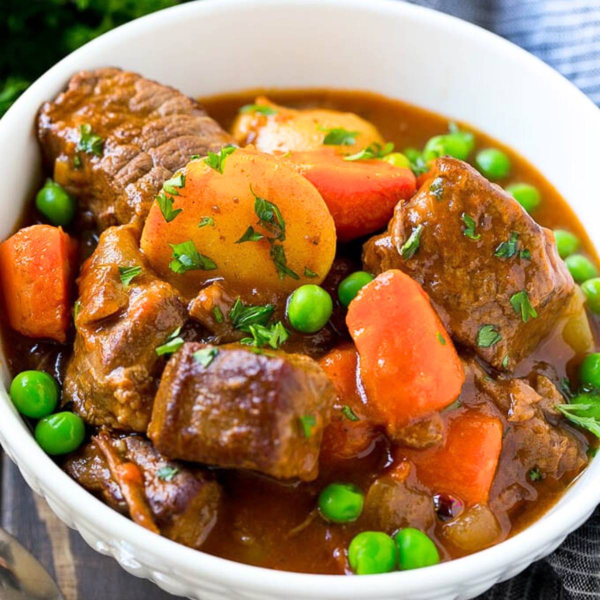 A white bowl filled with a hearty beef stew. The stew is made with tender chunks of beef, carrots, potatoes, and peas, all simmered in a rich, flavorful gravy.