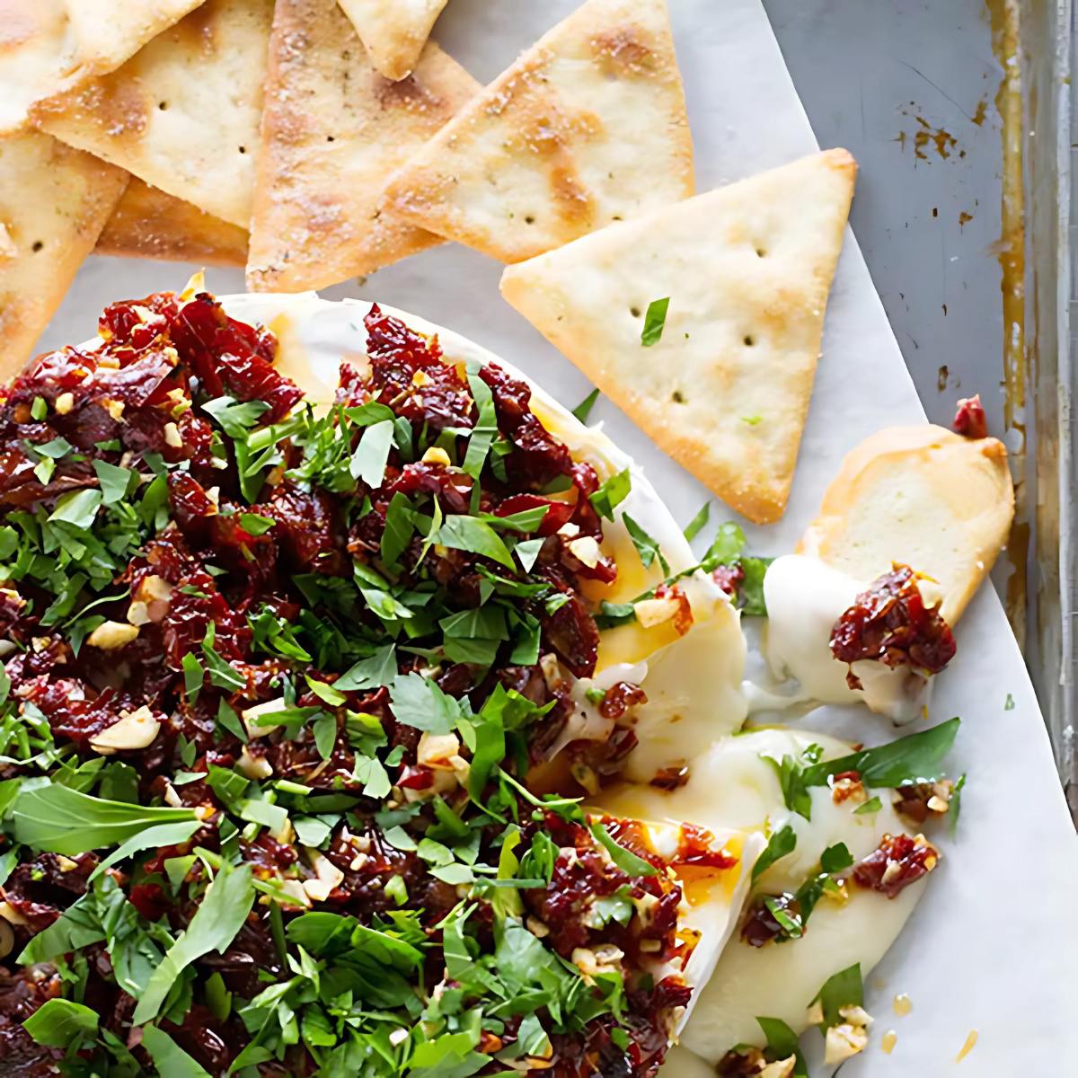 A wheel of baked brie topped with a mixture of sun-dried tomatoes, herbs, and nuts. Crackers surround the brie.