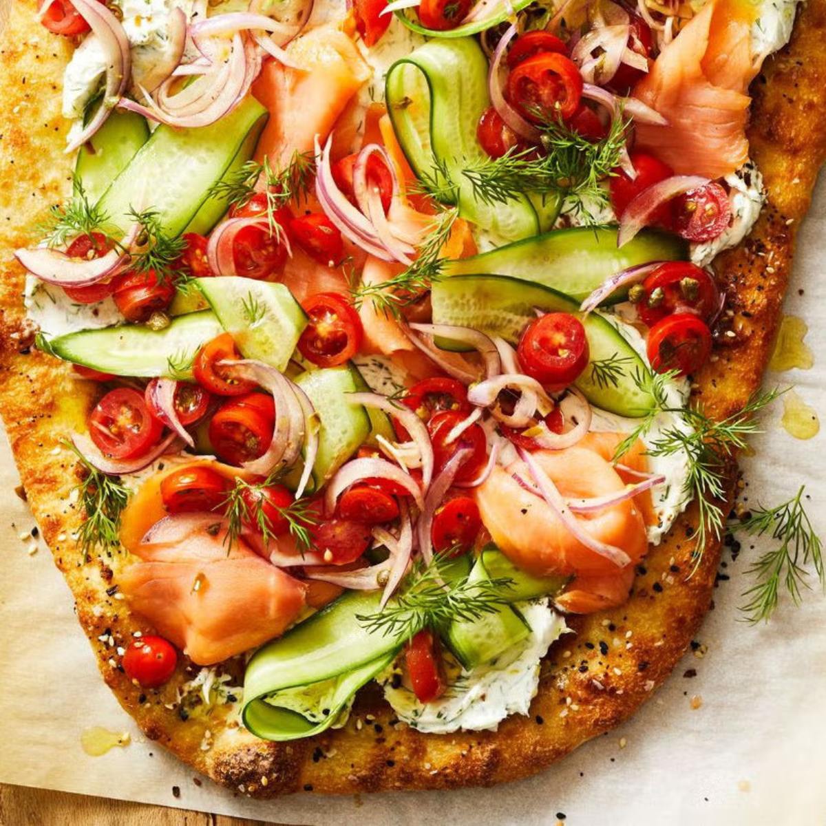 A flatbread pizza topped with smoked salmon, cucumbers, cherry tomatoes, red onion, dill, and cream cheese.