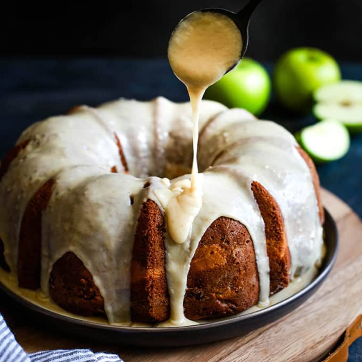 A golden-brown bundt cake drizzled with a creamy glaze, with a spoon pouring more glaze over the top.