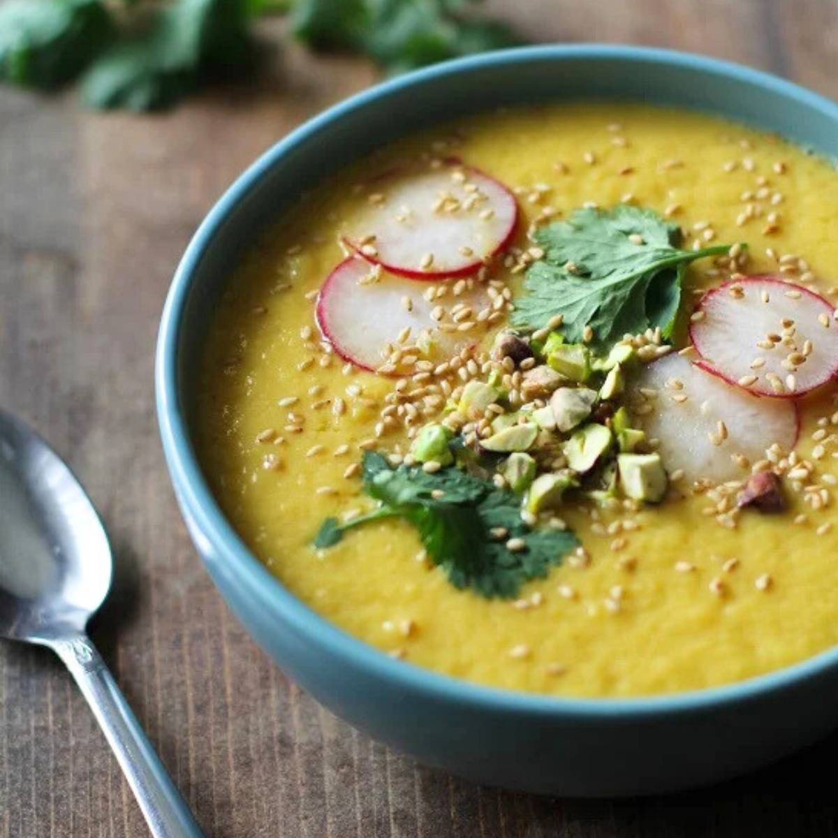 A bowl of golden beet soup topped with radish slices, cilantro, pistachios, and sesame seeds.