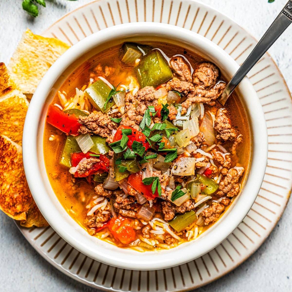 A white bowl filled with a flavorful soup with ground beef, rice, carrots, and bell peppers. The soup is topped with fresh parsley and is served with toasted bread.