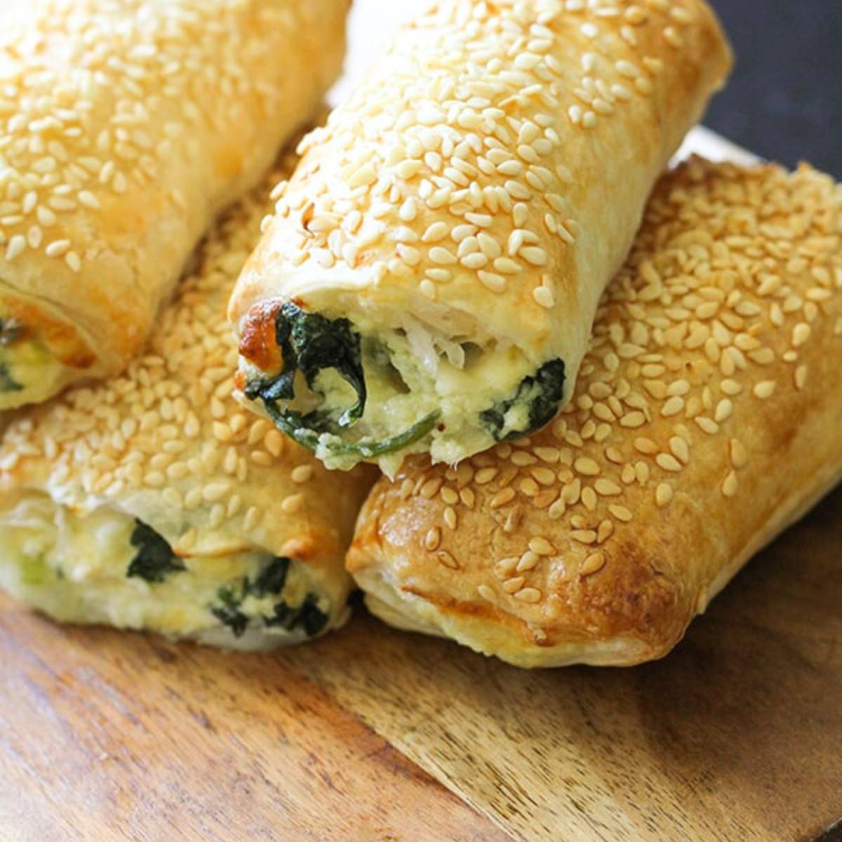 A close-up of a stack of golden-brown spinach and feta rolls sprinkled with sesame seeds.