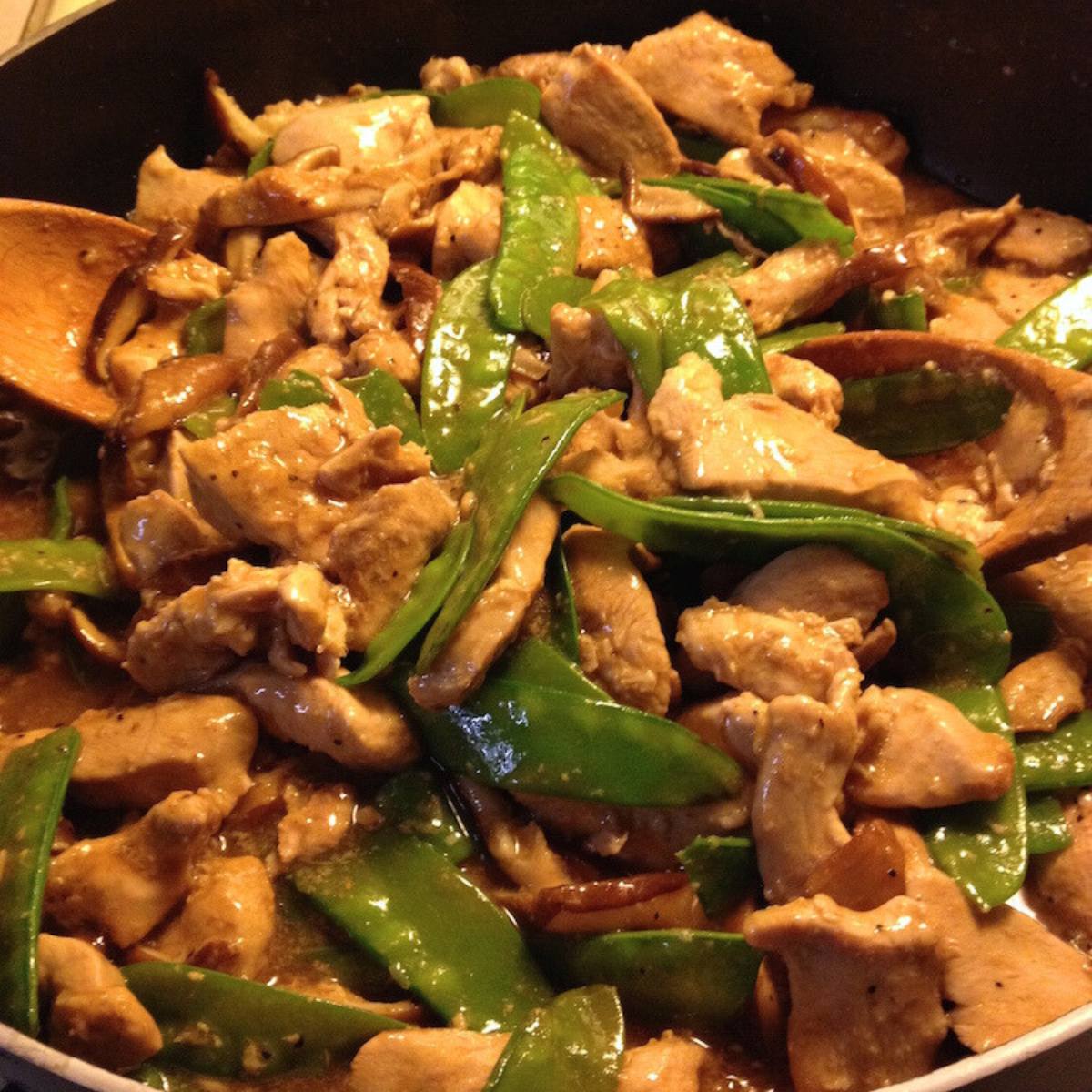 A skillet filled with a delicious-looking stir-fry. The stir-fry features tender pieces of pheasant, crisp snow peas, and sliced mushrooms, all coated in a savory sauce. A wooden spoon is resting in the pan.