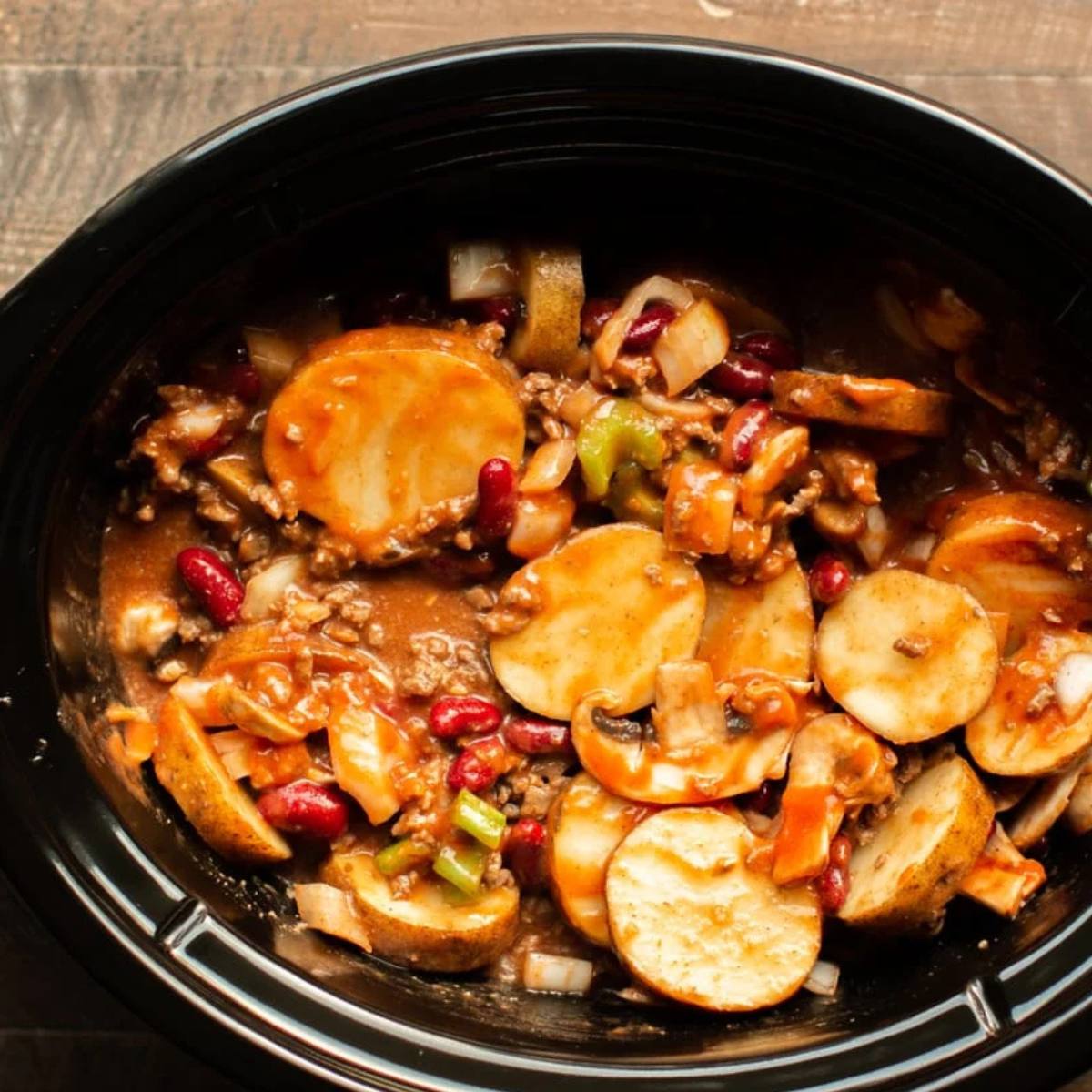 A slow cooker filled with ingredients for a hearty chili. The chili contains potatoes, beans, ground beef, onions, and other vegetables.