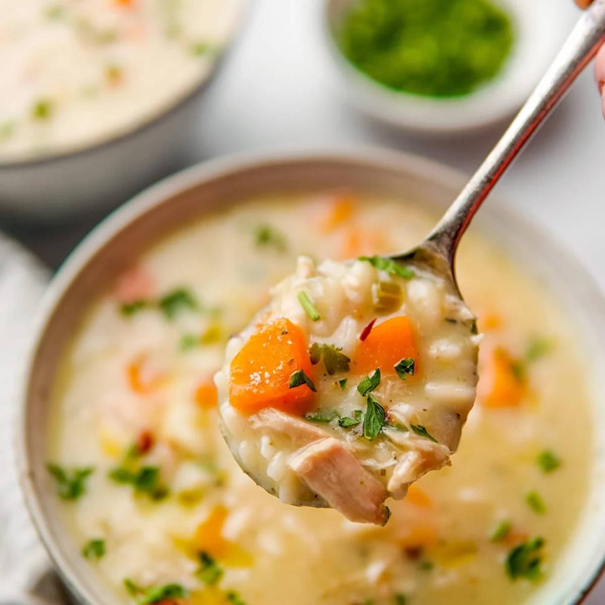 A spoon scooping out a spoonful of creamy chicken soup with rice, carrots, and herbs.