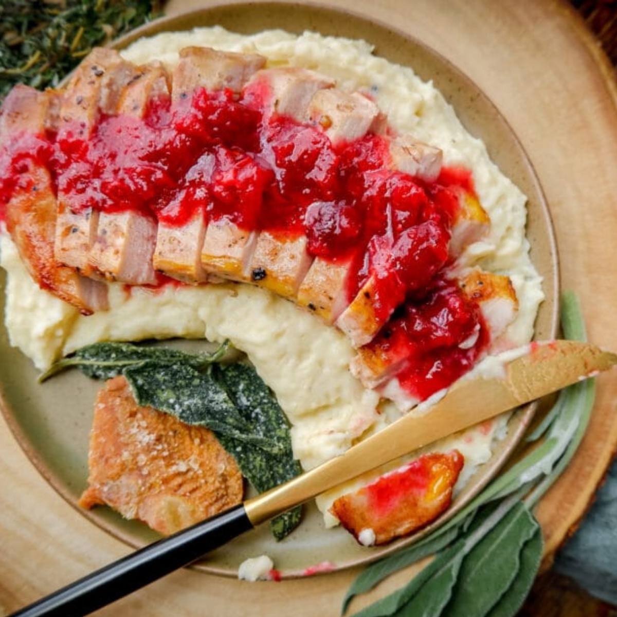 A delicious-looking plate of food. The plate features a pork chop topped with a rich, red cranberry sauce. The chopped pheasant is resting on a bed of creamy mashed potatoes. There's also a side of crispy bacon and a sprig of sage.