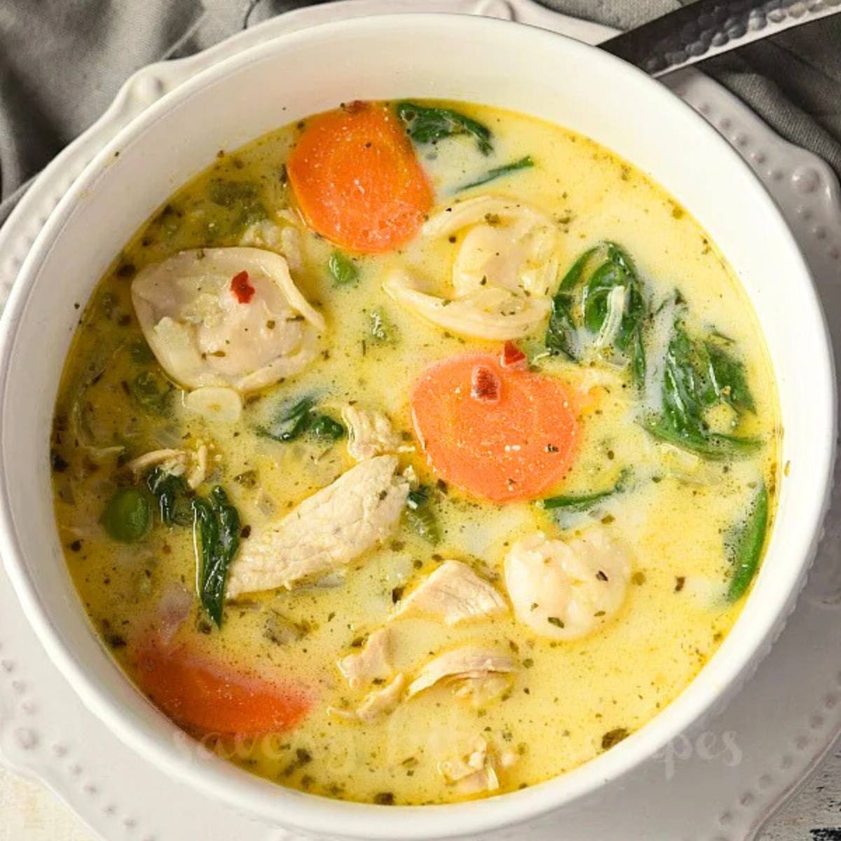 A bowl of creamy chicken and tortellini soup with vegetables. The soup is a light yellow color and is filled with tortellini, carrots, peas, spinach, and chicken.