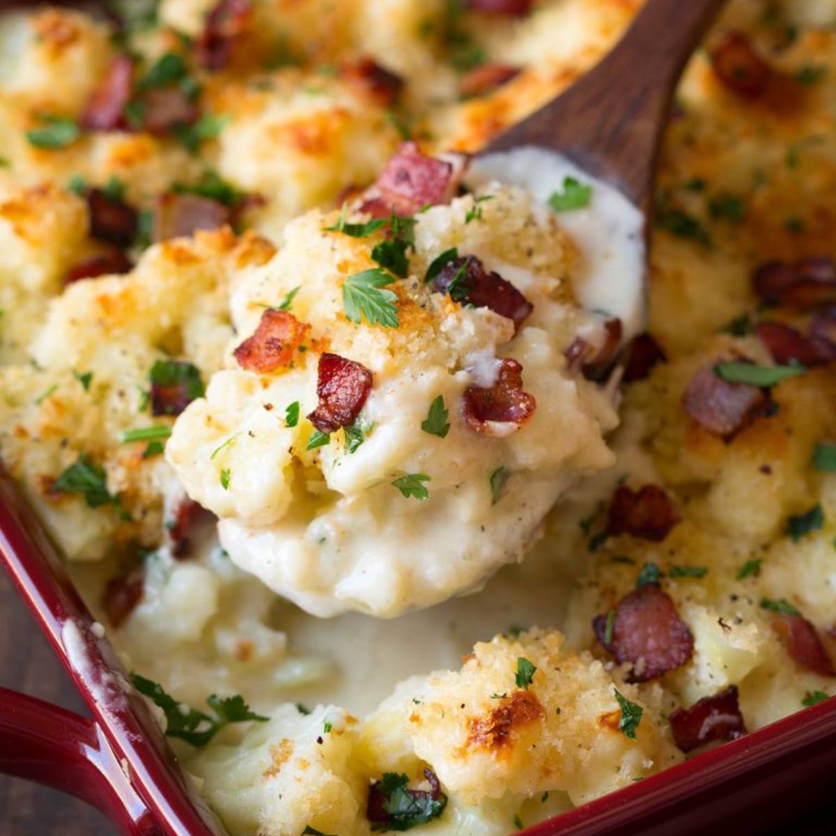 A close-up of a creamy cauliflower casserole with bacon and cheese, being scooped up with a wooden spoon.