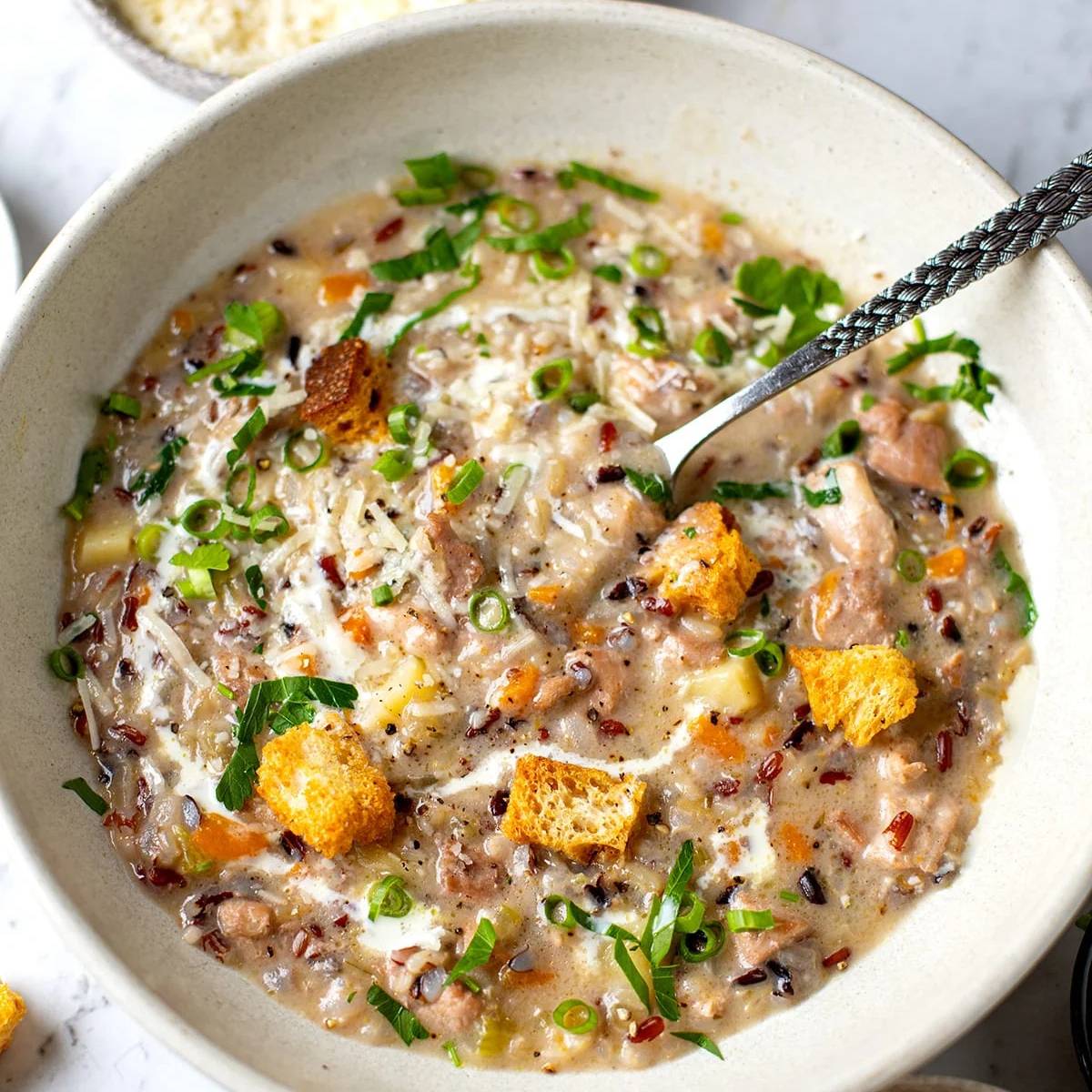 A close-up shot of a white bowl filled with a creamy wild rice soup, garnished with fresh herbs and croutons.