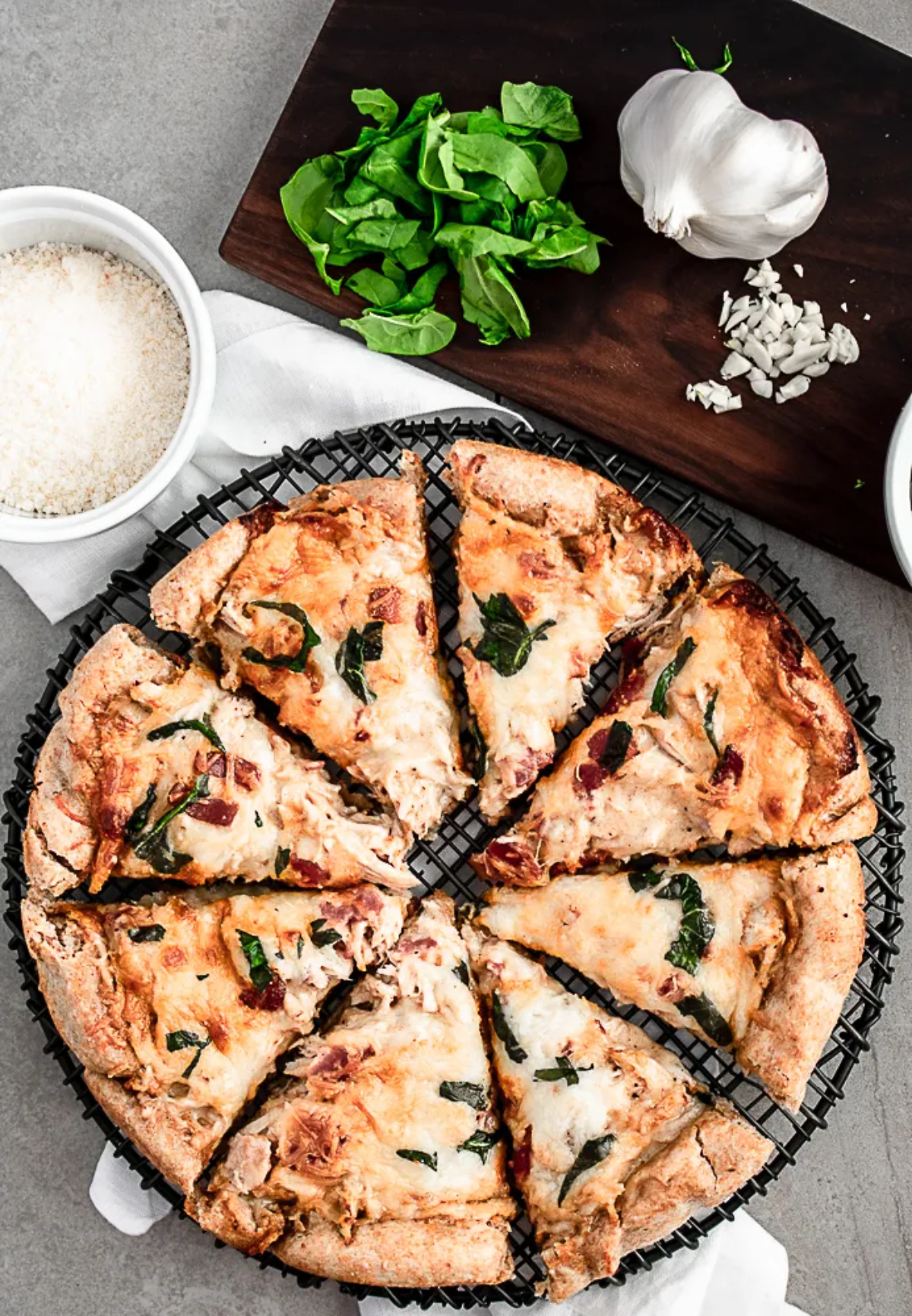 A delicious-looking pizza with a crispy crust and a variety of toppings. The pizza is cut into slices and arranged on a wire rack. There's a pot of white sauce and a bowl of shredded pheasant and cheese in the background.