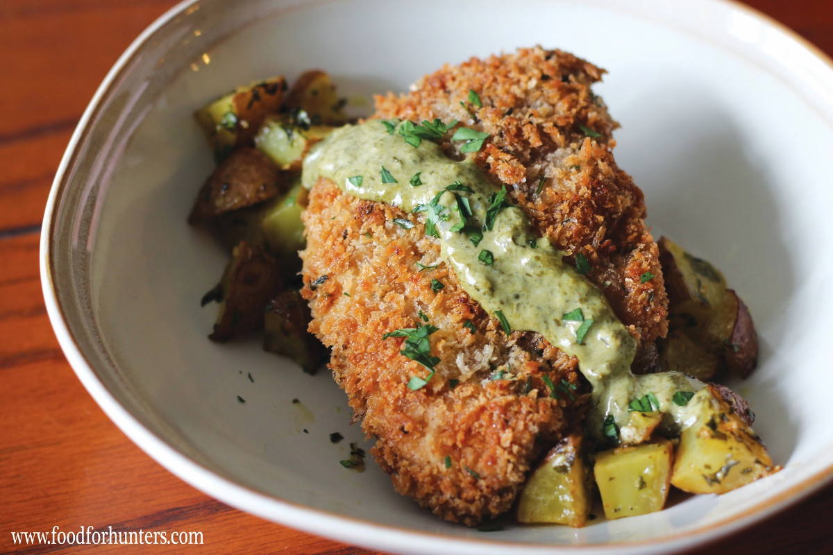 A golden-brown, breaded pheasant cutlet served with roasted potatoes and a creamy green sauce. The dish looks crispy and delicious.