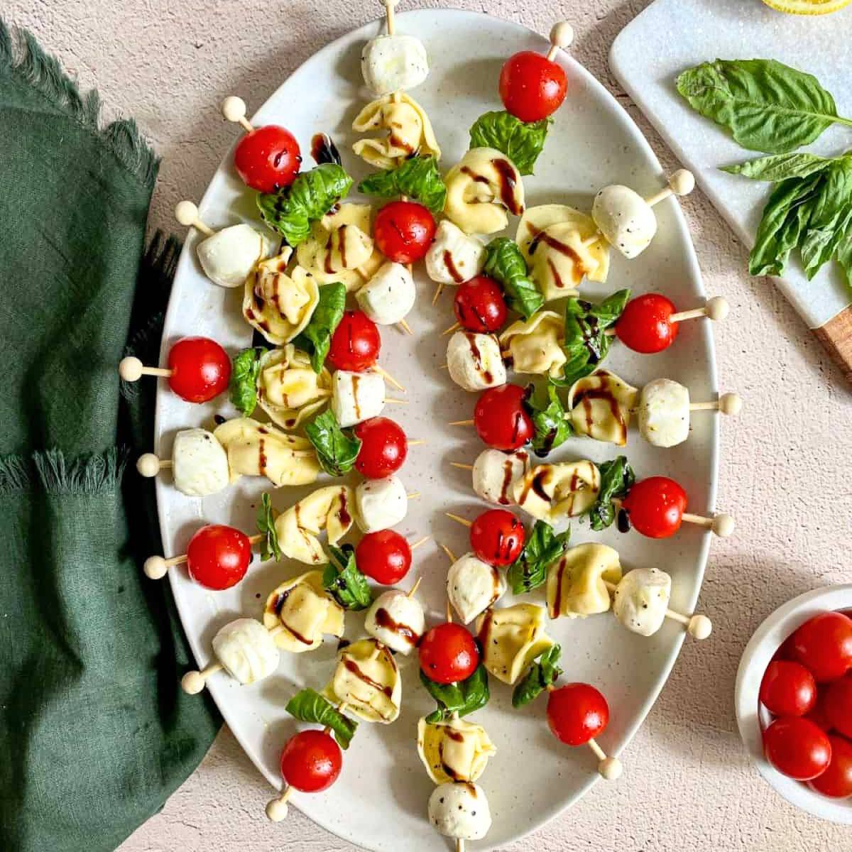 A platter of Caprese Tortellini Skewers with Balsamic Glaze. The skewers are arranged in a circular pattern and feature alternating pieces of tortellini, cherry tomatoes, fresh mozzarella, and basil leaves. 