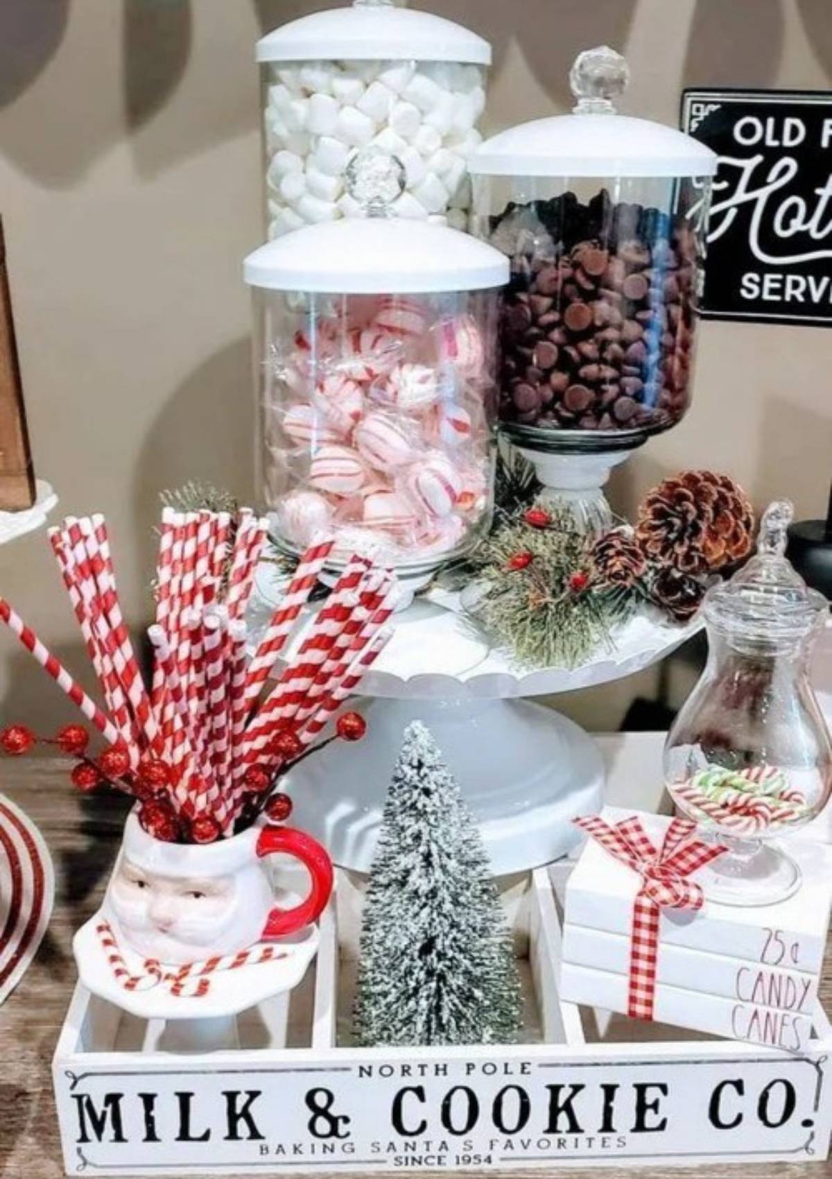 A festive hot cocoa bar with jars of marshmallows, chocolate chips, and candy canes. There is also a small Christmas tree, a Santa Claus mug, and a sign that says "Milk & Cookie Co. Baking Santa's Favorites Since 1954".