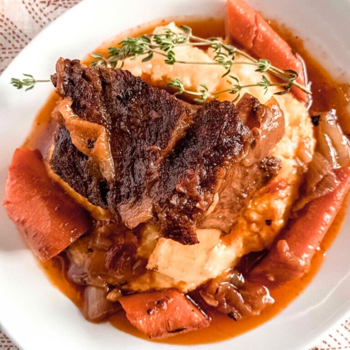 A hearty plate of braised short ribs served over creamy polenta. The rich, tender meat is cooked in a flavorful sauce with carrots and onions, and garnished with fresh thyme.