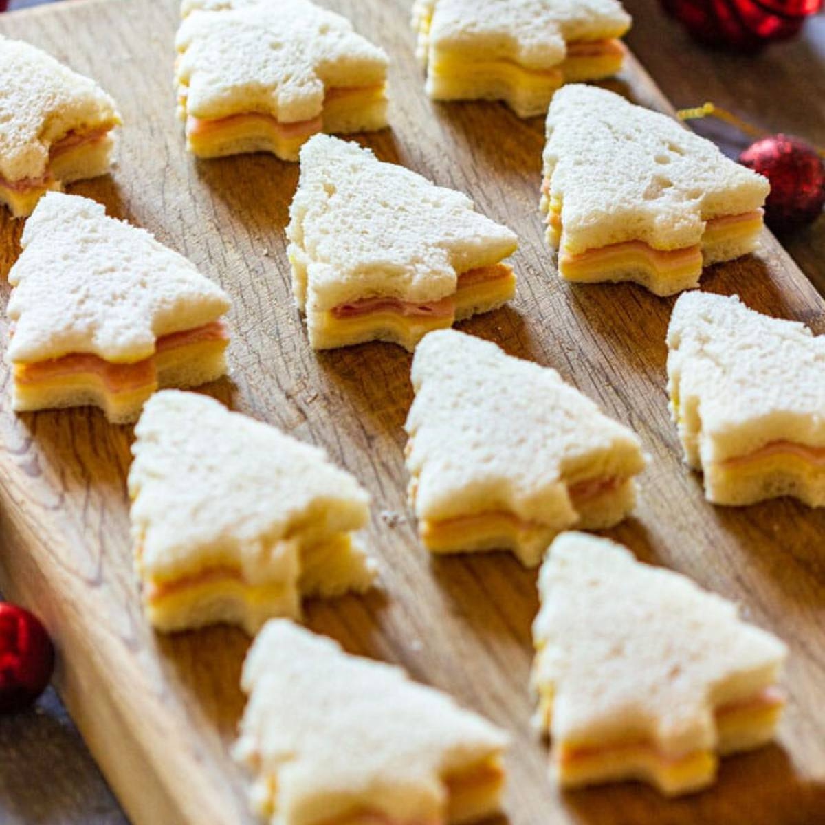 A wooden board filled with bite-sized Christmas tree sandwiches. The sandwiches are made with white bread, ham, and cheese, and are cut into the shape of Christmas trees.