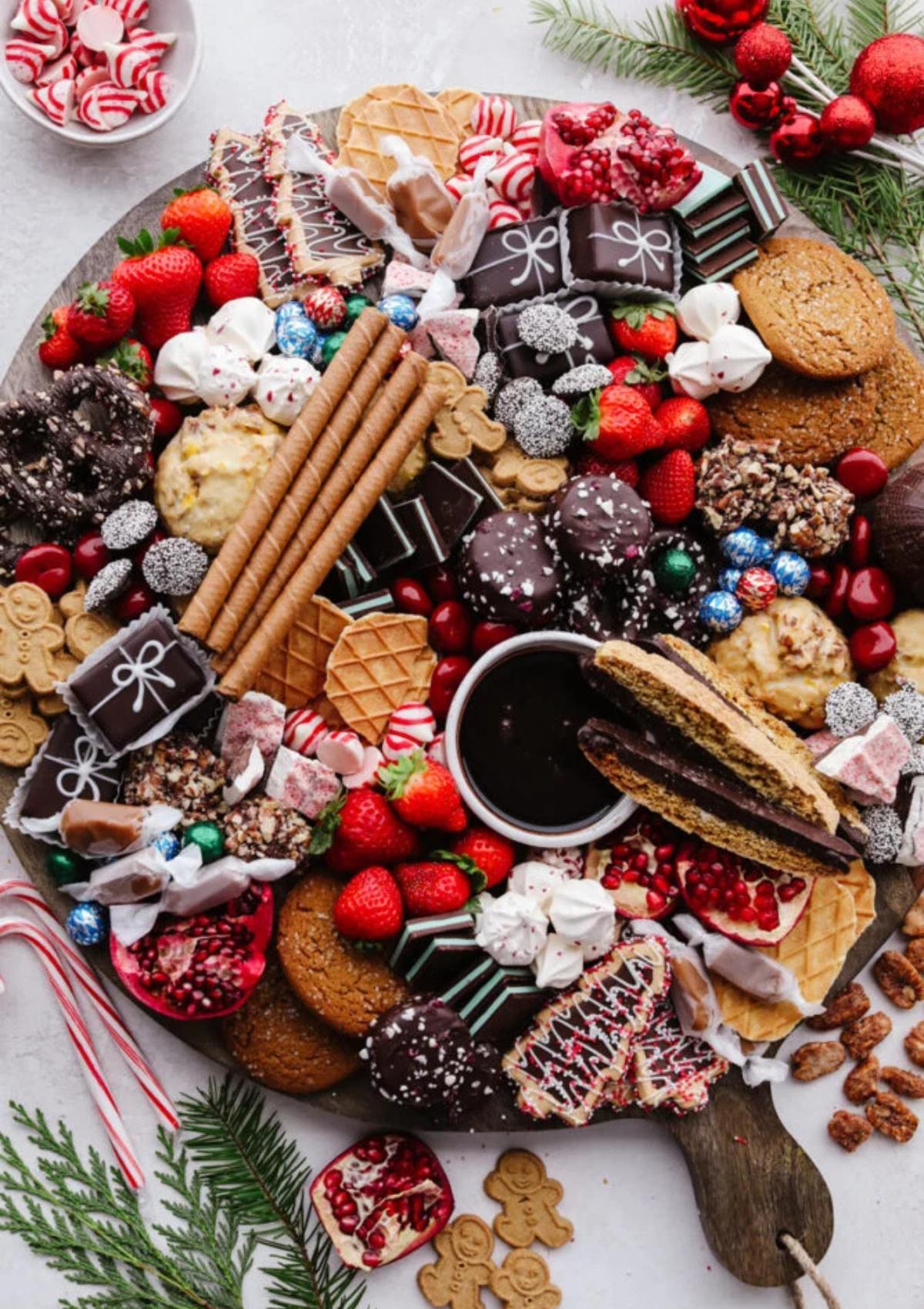 A festive Christmas dessert charcuterie board filled with an assortment of delicious treats. The board includes cookies, chocolates, candies, fruits, and a small bowl of dipping sauce.