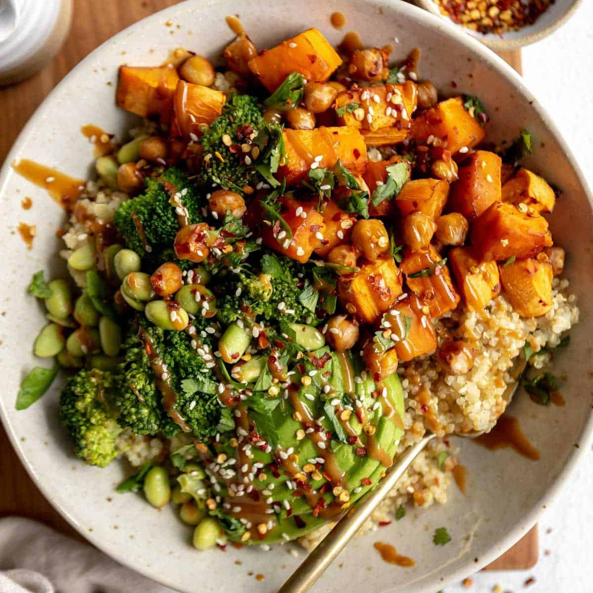 A colorful Buddha bowl with quinoa, roasted sweet potatoes, chickpeas, broccoli, edamame, avocado, and a drizzle of sauce.