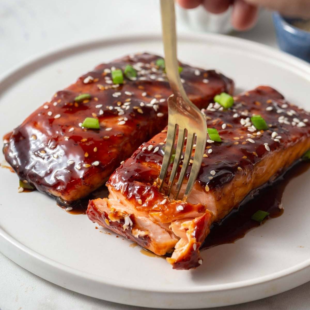 The salmon fillets are glazed with a sticky, teriyaki sauce and garnished with sesame seeds and green onions. A fork is being used to flake the salmon, which looks perfectly cooked and tender.