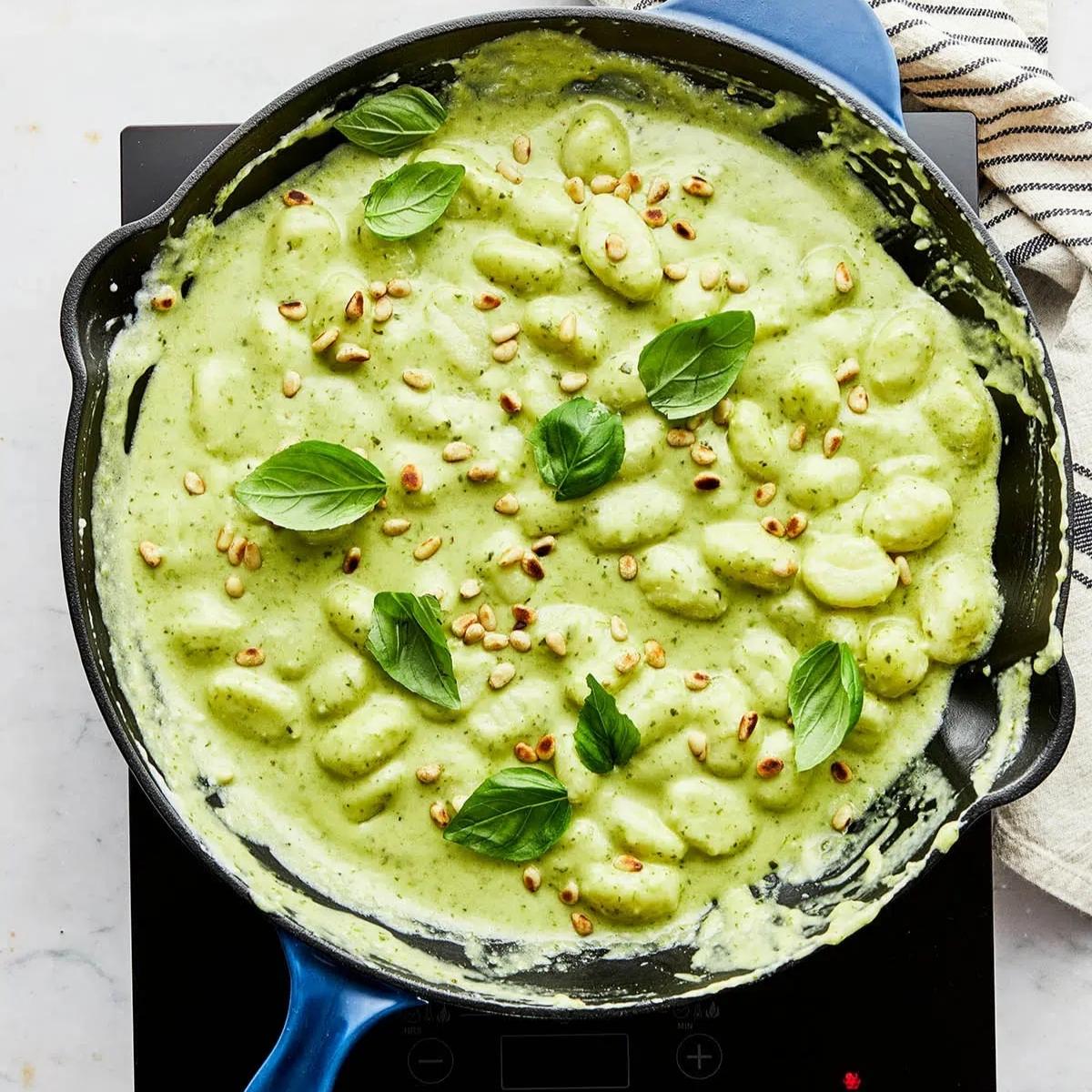 A cast iron skillet filled with creamy pesto gnocchi. The gnocchi is coated in a vibrant green pesto sauce and garnished with fresh basil leaves and toasted pine nuts.