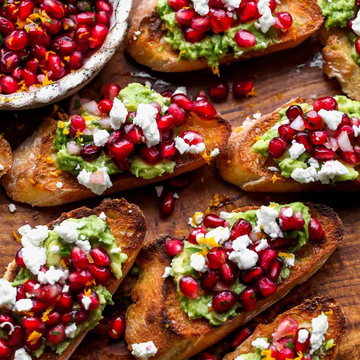 A wooden board filled with slices of toasted bread topped with mashed avocado, feta cheese, pomegranate seeds, and herbs.