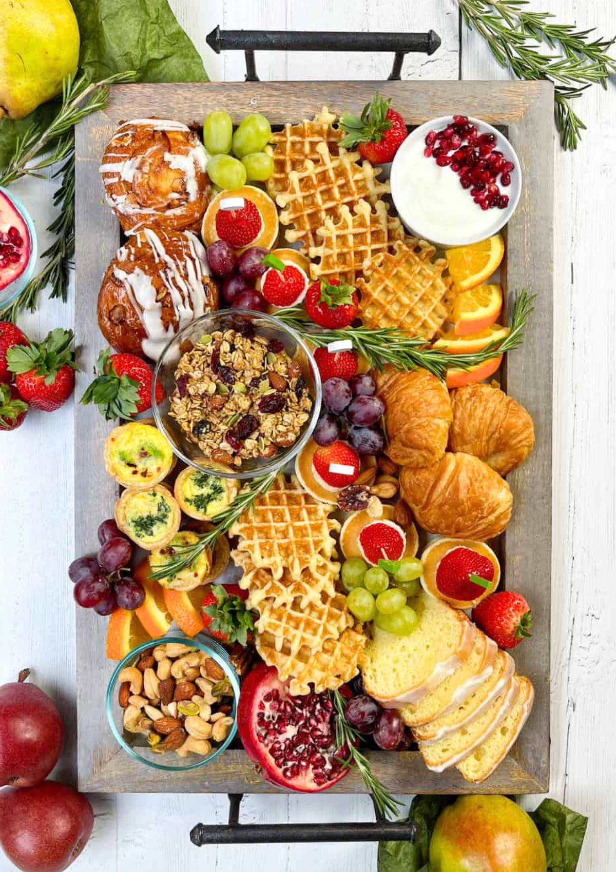 A festive breakfast charcuterie board filled with a variety of delicious treats. The board includes pastries, fruits, yogurt, granola, nuts, and waffles.