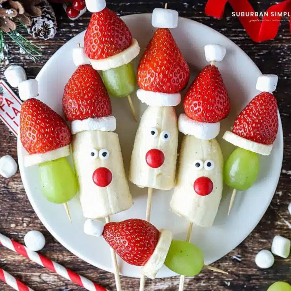 A plate of festive fruit skewers featuring bananas, strawberries, and grapes. Some of the skewers are decorated with marshmallows to resemble Santa hats, while others have googly eyes and red candies to look like reindeer faces.