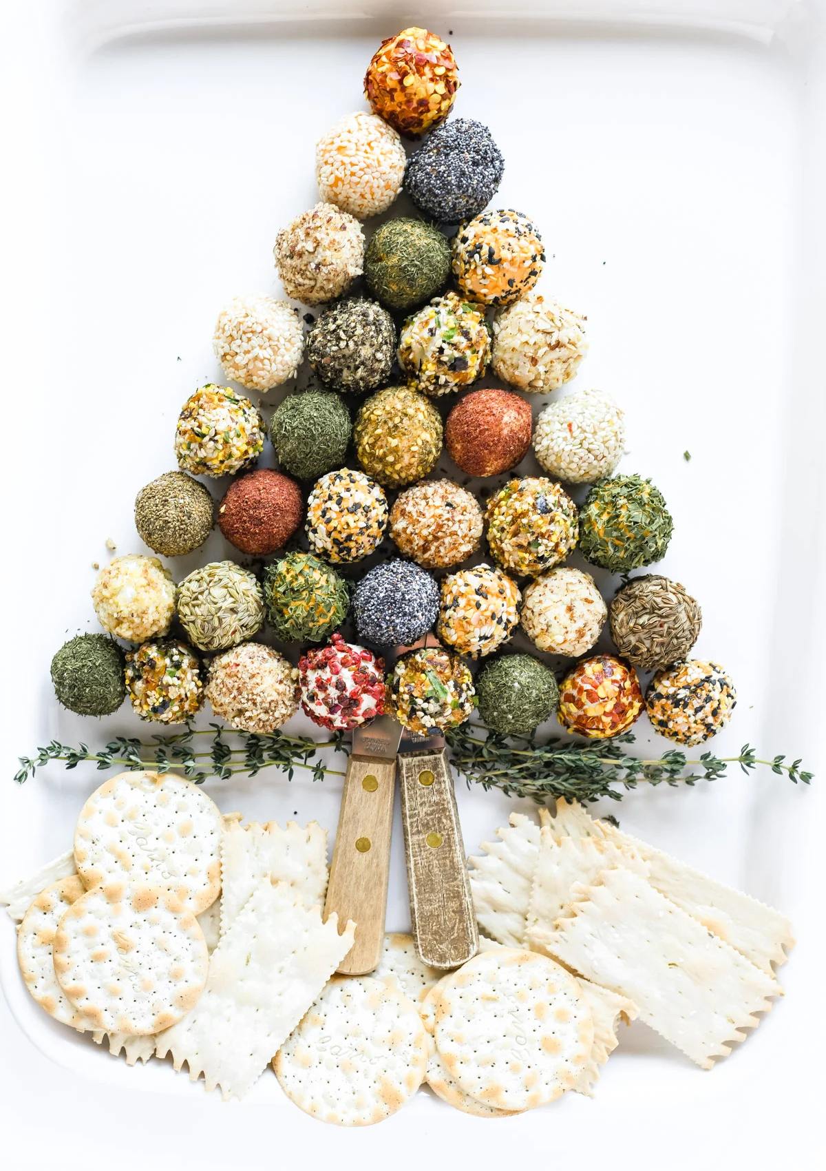 A festive Christmas tree-shaped platter of colorful cheese balls, arranged in a triangular pattern. The cheese balls are coated in various herbs, spices, and seeds, and surrounded by crackers and a sprig of thyme.