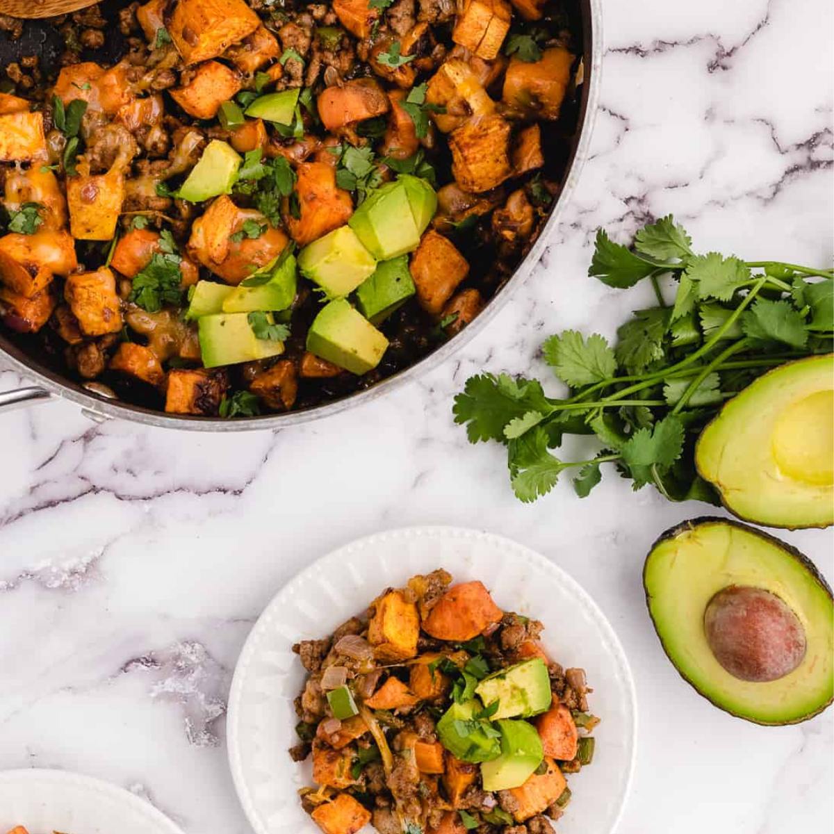 A skillet filled with a savory mixture of ground beef, sweet potatoes, peppers, and cheese, topped with avocado slices and cilantro.