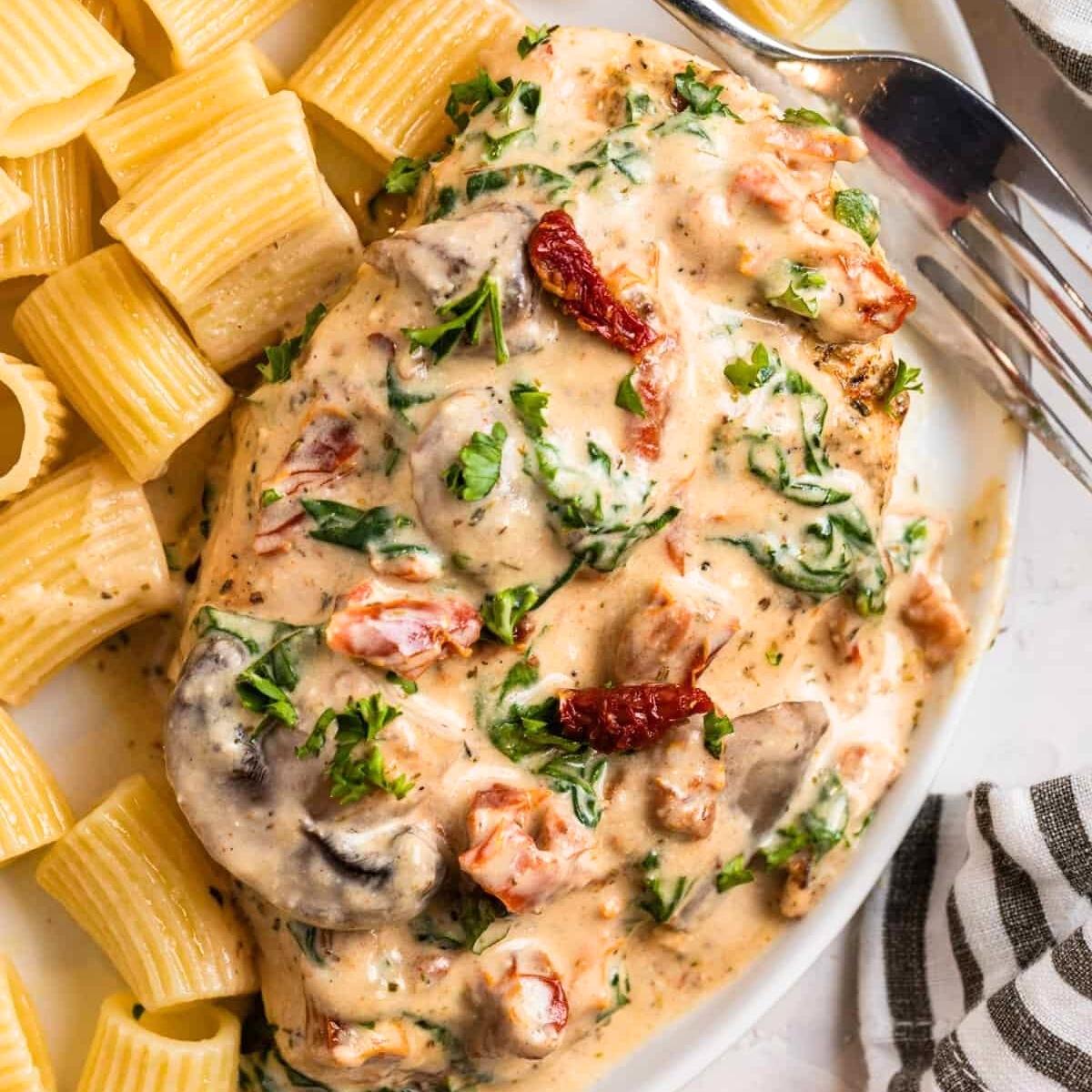 A plate of creamy Tuscan chicken with sun-dried tomatoes and mushrooms, served over a bed of cooked pasta. The dish is garnished with fresh parsley.