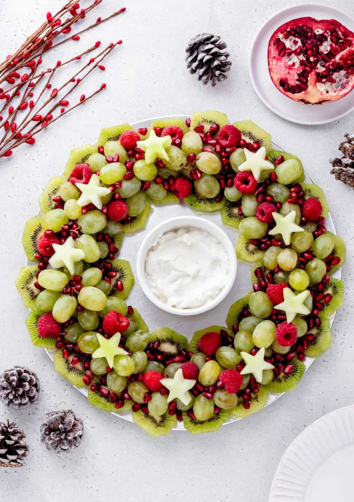 A festive fruit wreath made with grapes, kiwi, raspberries, pomegranate seeds, and star-shaped melon slices. A small bowl of dip is placed in the center.