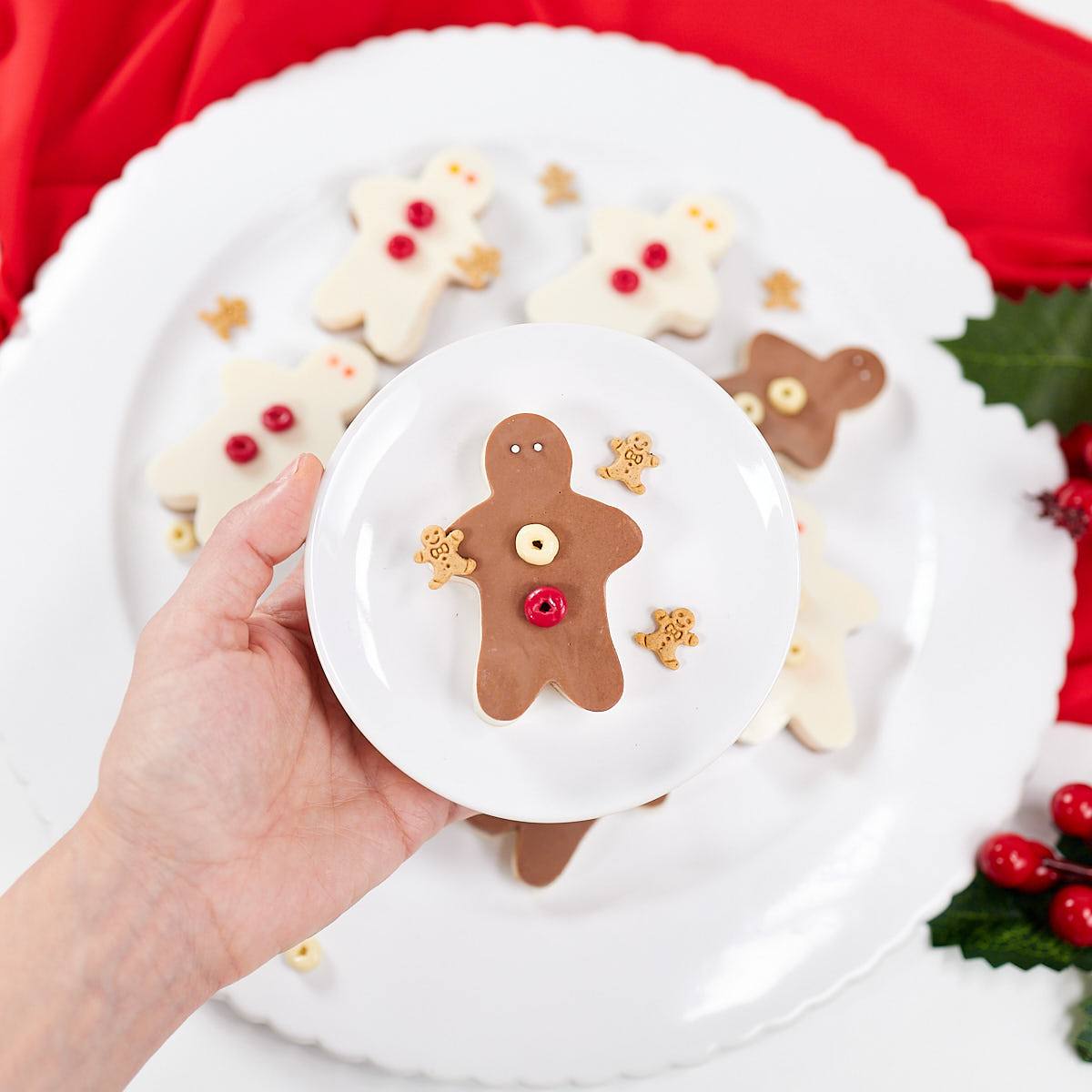 A hand holding a small plate with a gingerbread man made out of cheese. The gingerbread man is decorated with red and yellow candies for buttons and eyes. There are other cheese snacks shaped like gingerbread men on the plate and in the background.