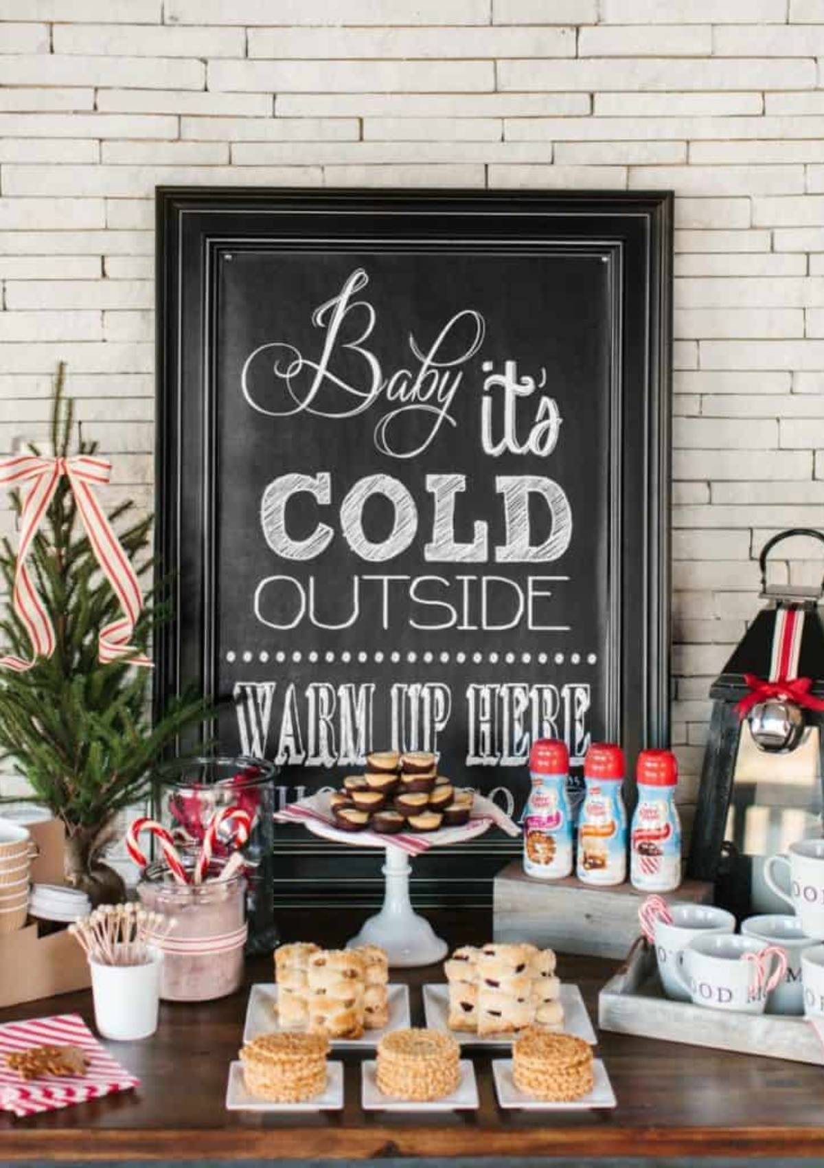 A festive hot cocoa bar with a chalkboard sign that says "Baby it's Cold Outside, Warm Up Here." The bar includes hot chocolate, marshmallows, candy canes, and cookies.