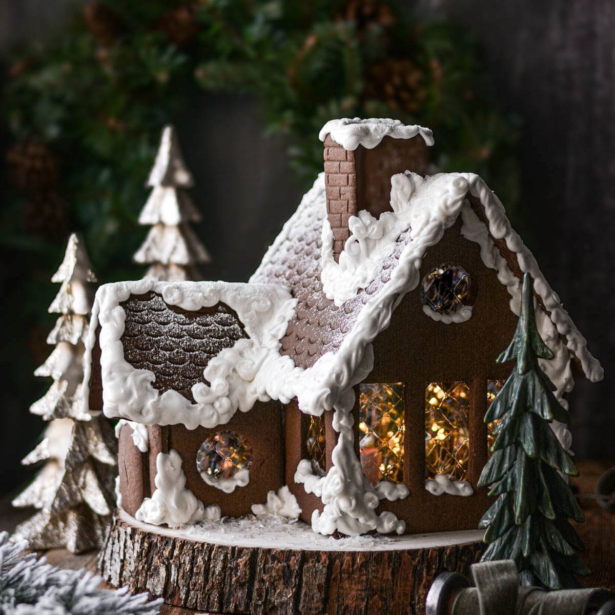 A festive gingerbread house with lit windows, nestled amongst small Christmas trees.