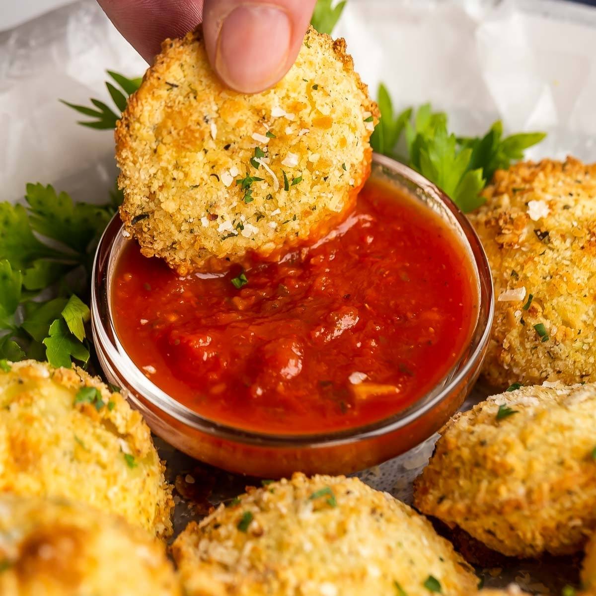 A hand dipping a crispy, golden-brown ravioli into a bowl of marinara sauce. The ravioli is breaded and looks delicious.