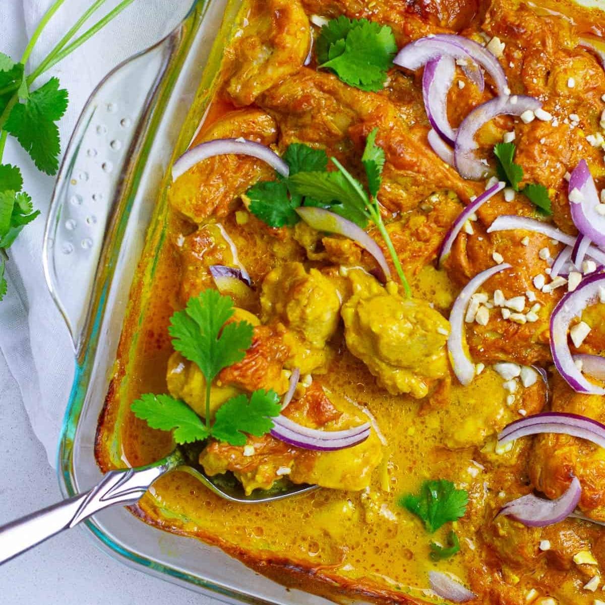 A close-up of a baking dish filled with a rich and creamy chicken curry. The chicken pieces are coated in a golden sauce, and the dish is garnished with chopped cilantro, sliced red onions, and chopped peanuts.