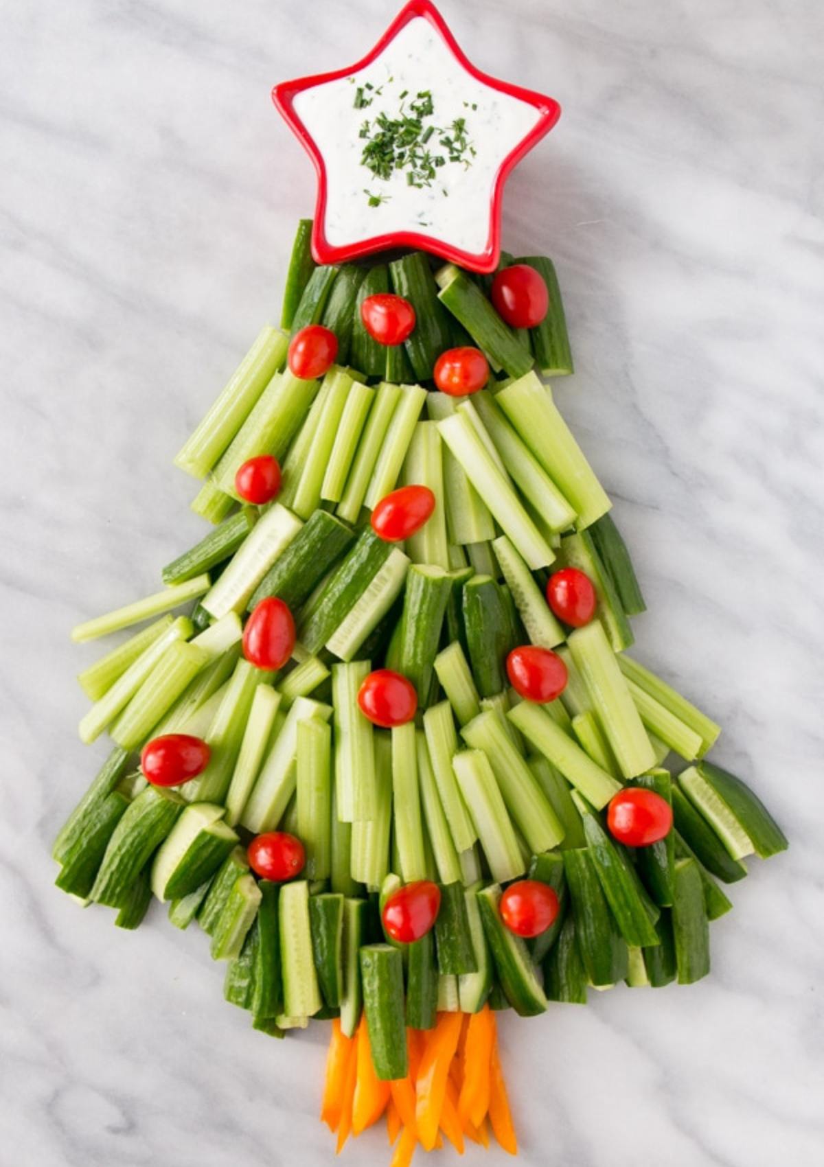 A festive Christmas tree-shaped vegetable platter made with celery, cucumber, cherry tomatoes, and carrots. A small bowl of dip is placed at the top to represent the star.