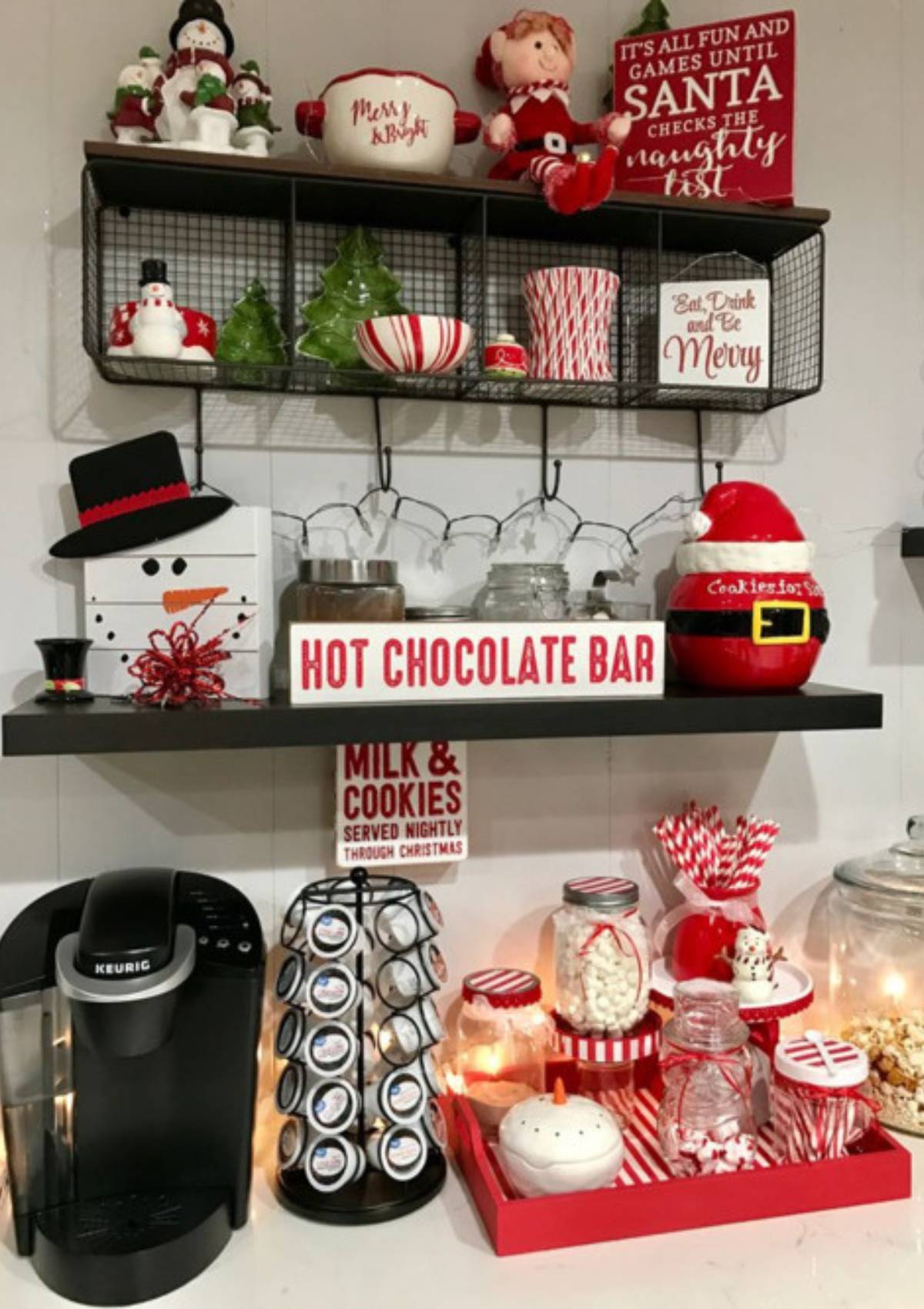 A festive hot chocolate bar with a Keurig coffee maker, jars of marshmallows and candy canes, and a sign that says "Hot Chocolate Bar." The bar is decorated with Christmas-themed decorations like snowmen and Santa Claus.