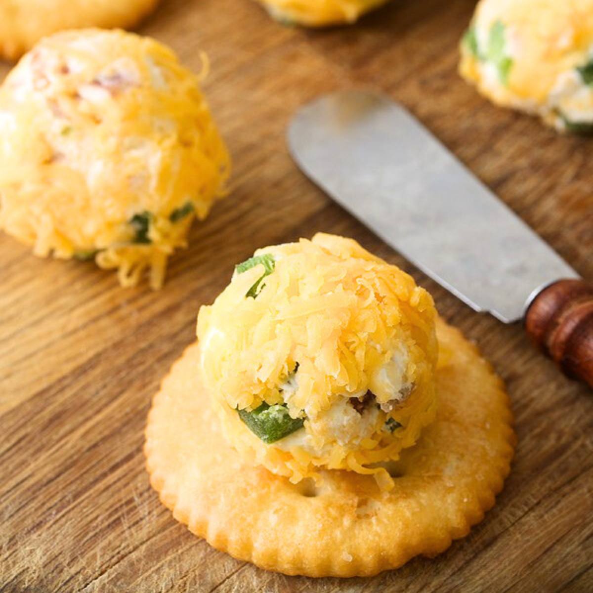 A cracker topped with a ball of cheese, bacon, and chives, coated in shredded cheddar cheese. A butter knife is nearby.








