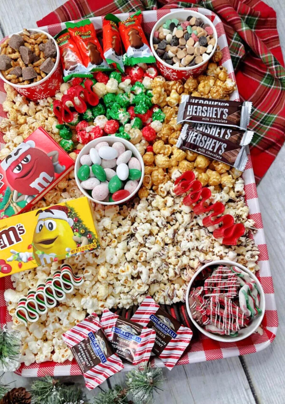 A festive Christmas snack board filled with popcorn, candy, and chocolate. The board is decorated with Christmas-themed treats like candy canes and red and green sprinkles.