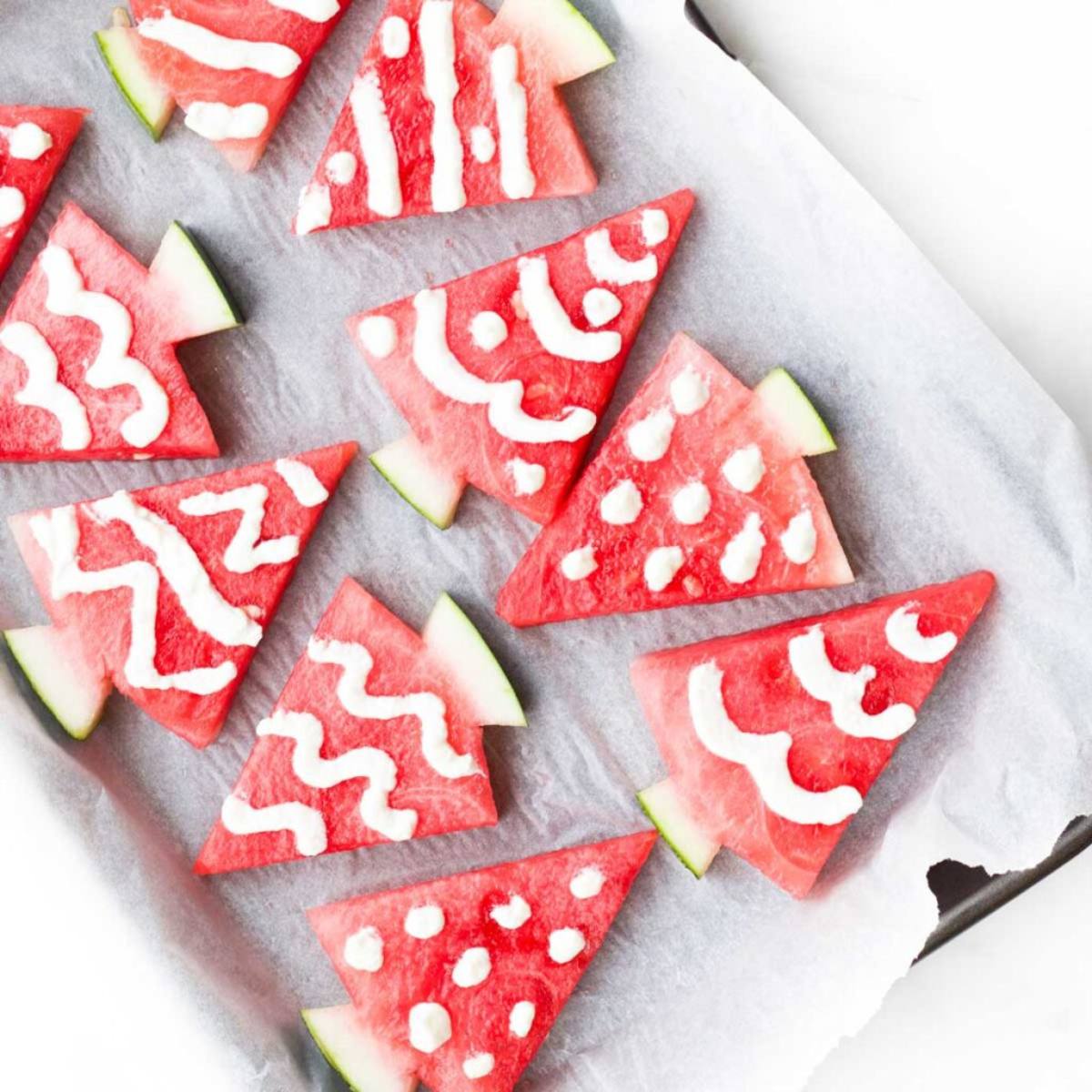 A baking sheet filled with festive watermelon Christmas trees. The watermelon slices are cut into tree shapes and decorated with white frosting to resemble snow and ornaments.
